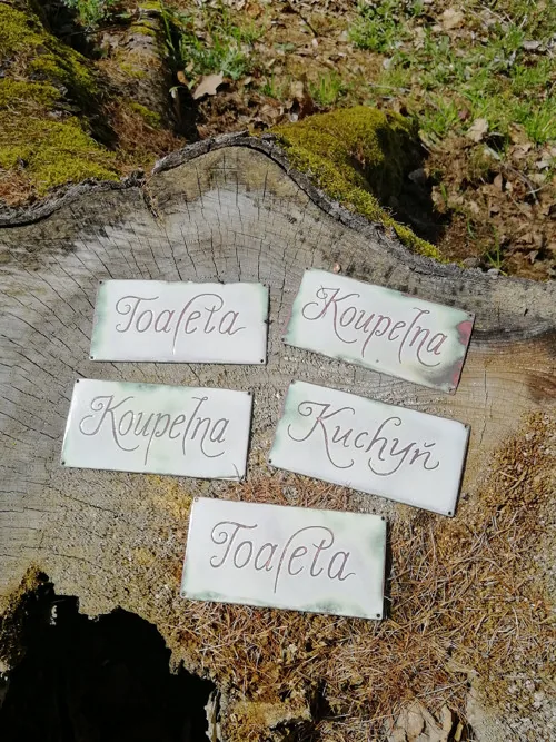Gesamtansicht des Satzes: fünf kleine Emaille-Türschilder mit den Aufschriften „Toaleta“, „Koupelna“ und „Kuchyň“, ausgelegt auf einem Baumstumpf.